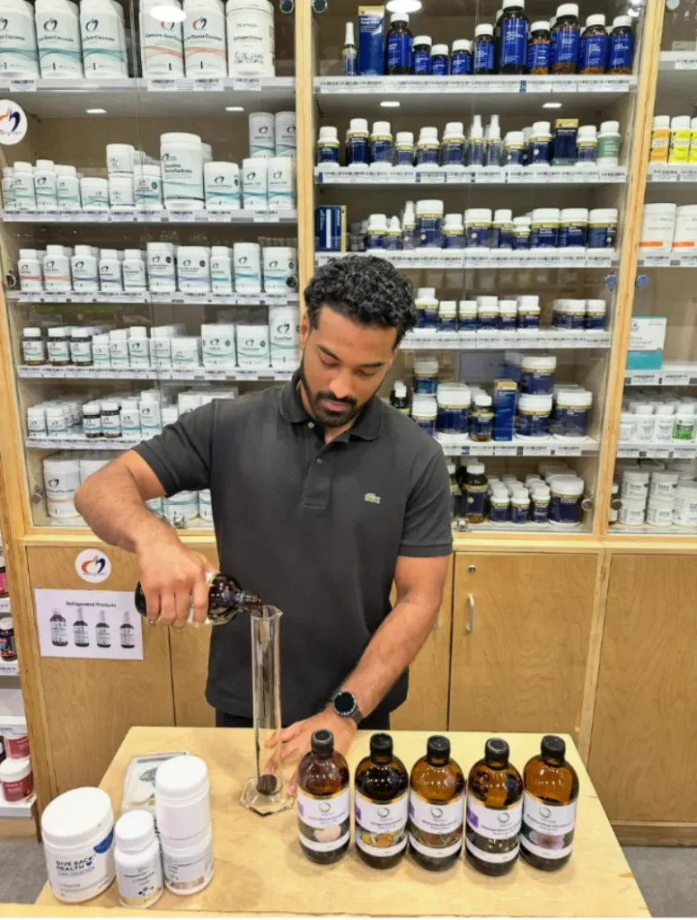 Naturopath preparing a liquid herbal formulation in front of shelves stocked with practitioner-grade supplements and natural health products.